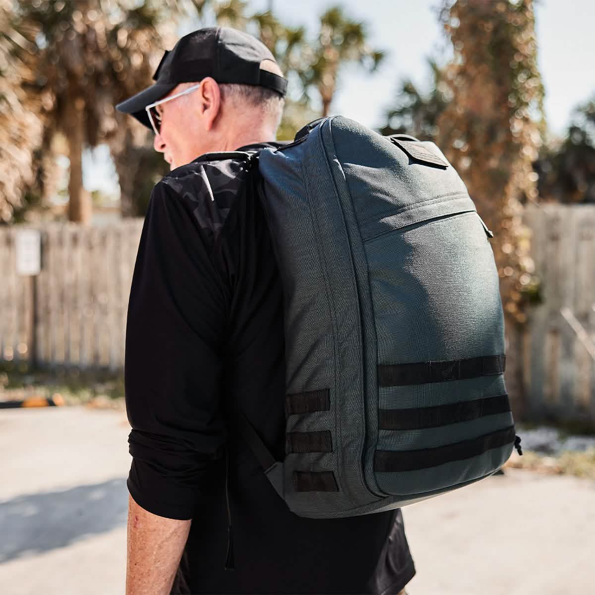 A man wearing a black cap and sunglasses carries a large gray backpack, similar to the rugged GR1 USA - Cordura (The Original Ruck) by GORUCK. He walks outdoors on a sunny day, with a wooden fence and trees in the background, embodying the spirit of Special Forces endurance.