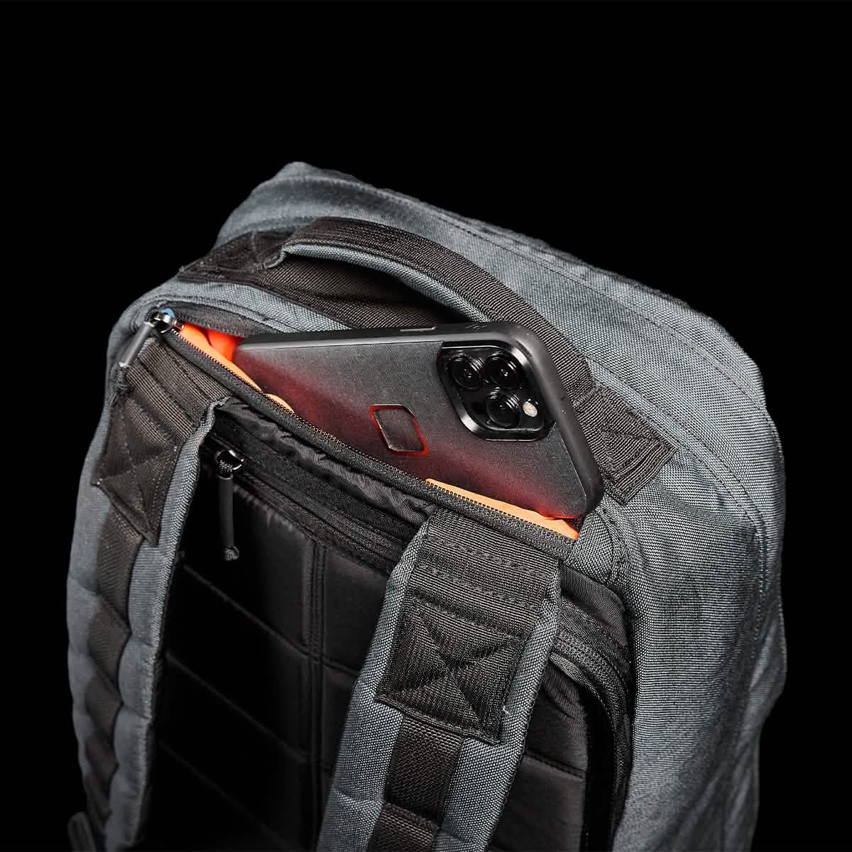 A smartphone peeks from the top pocket of a dark-colored backpack with padded straps, highlighting its GR1 USA - Cordura design by GORUCK and bombproof laptop compartment, set against a black background.