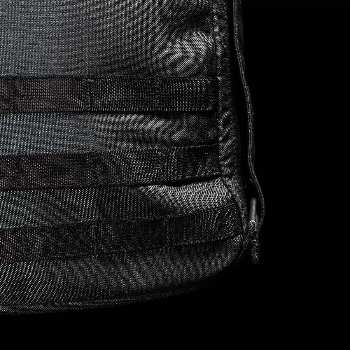 A close-up of the GORUCK GR1 USA - Cordura showcases its black tactical design, featuring horizontal nylon webbing straps and a zippered edge on the right. The fabric's texture and stitching details highlight the rugged resilience perfect for a GORUCK Challenge, set against a plain black background.
