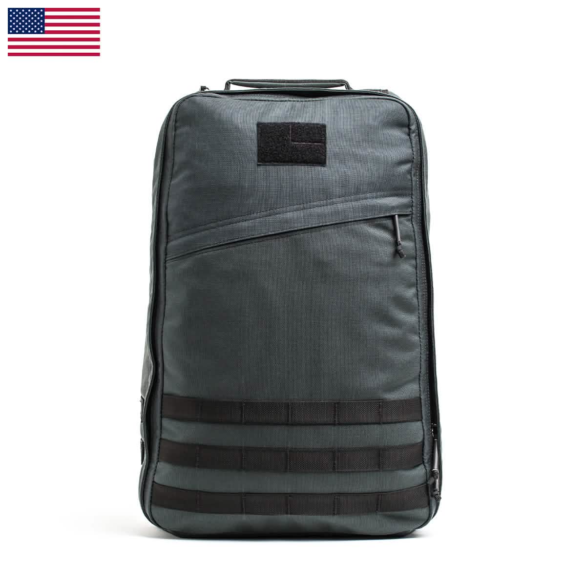 The GR1 USA - Cordura (The Original Ruck) by GORUCK, featuring a gray tactical design emblematic of American manufacturing, boasts a simplistic style with multiple zippers and a horizontal strap pattern at the bottom. An American flag adorns the top left corner of the image, symbolizing its journey from Baghdad to NYC.