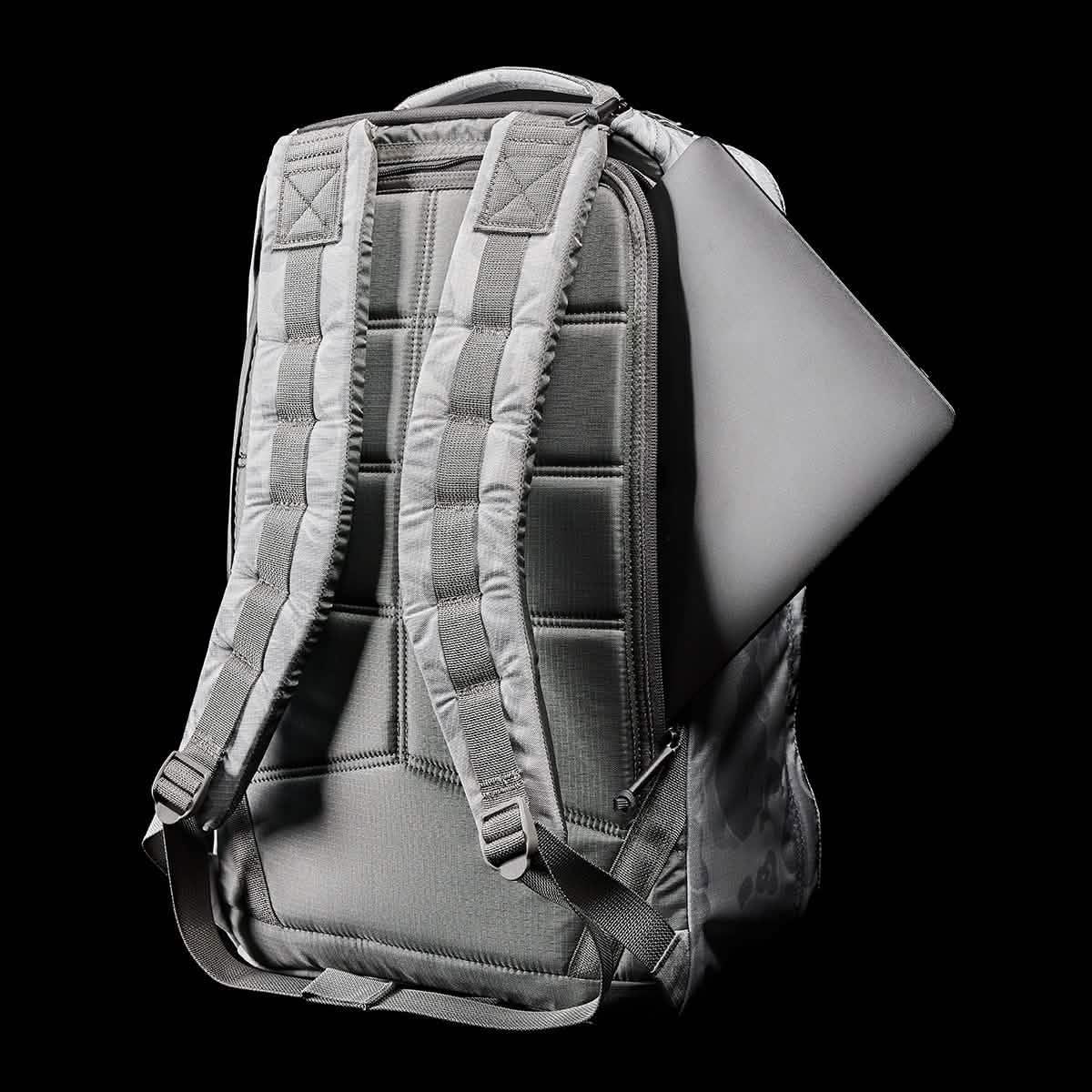 Back view of gray GORUCK rucksack with padded straps and a laptop partially inserted in side compartment on black background