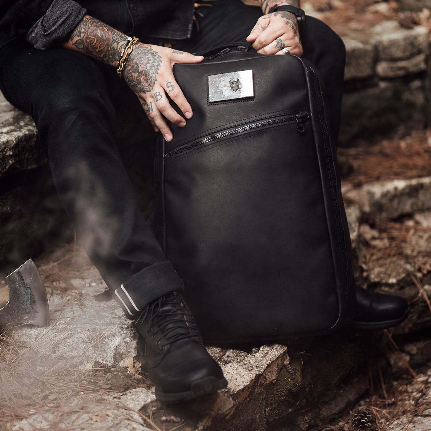 A tattooed person in black clothes sits on a stone ledge outdoors, holding the GR1 USA x Carryology - Berserker Viking backpack, evoking the fearless spirit of Viking warriors.