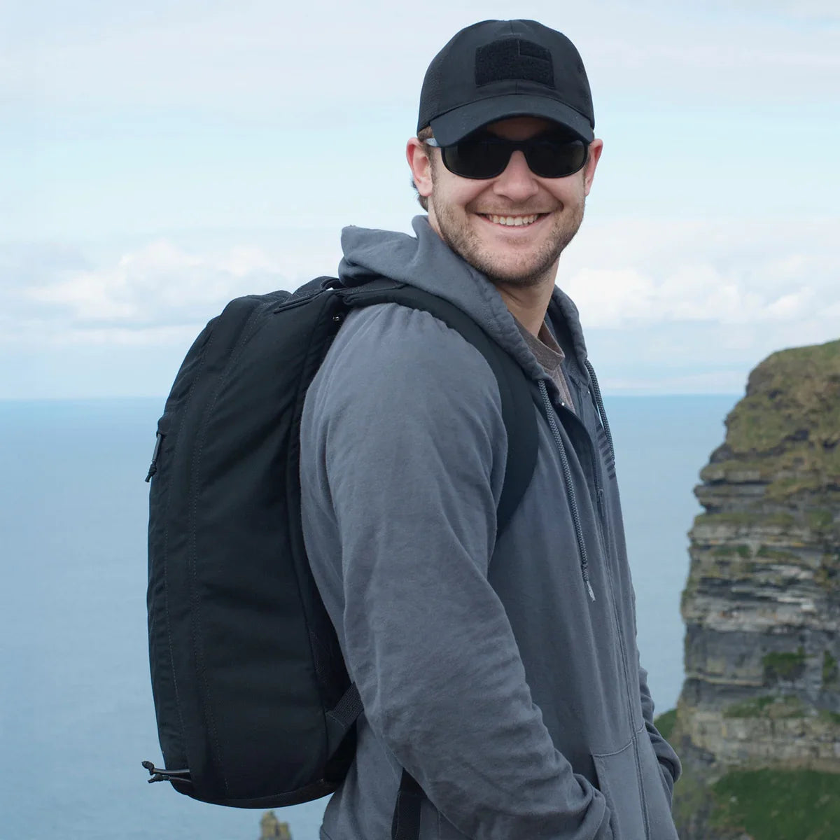 A man in a black cap and sunglasses, wearing a GR1 USA Slick - Ballistic Nylon Cordura backpack, smiles outdoors by a cliff with the sea behind him.