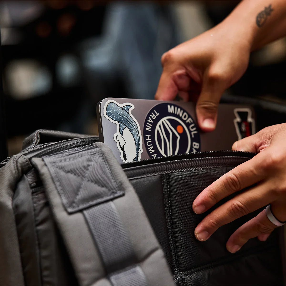 Person placing a sticker-covered laptop into a grey tactical GORUCK backpack pocket
