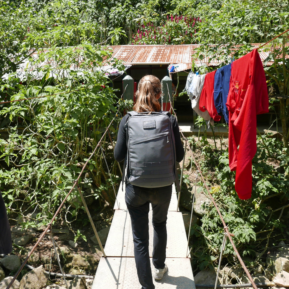 With the GR1 USA - Cordura everyday carry backpack, a person crosses a narrow bridge toward a house where laundry hangs outside.