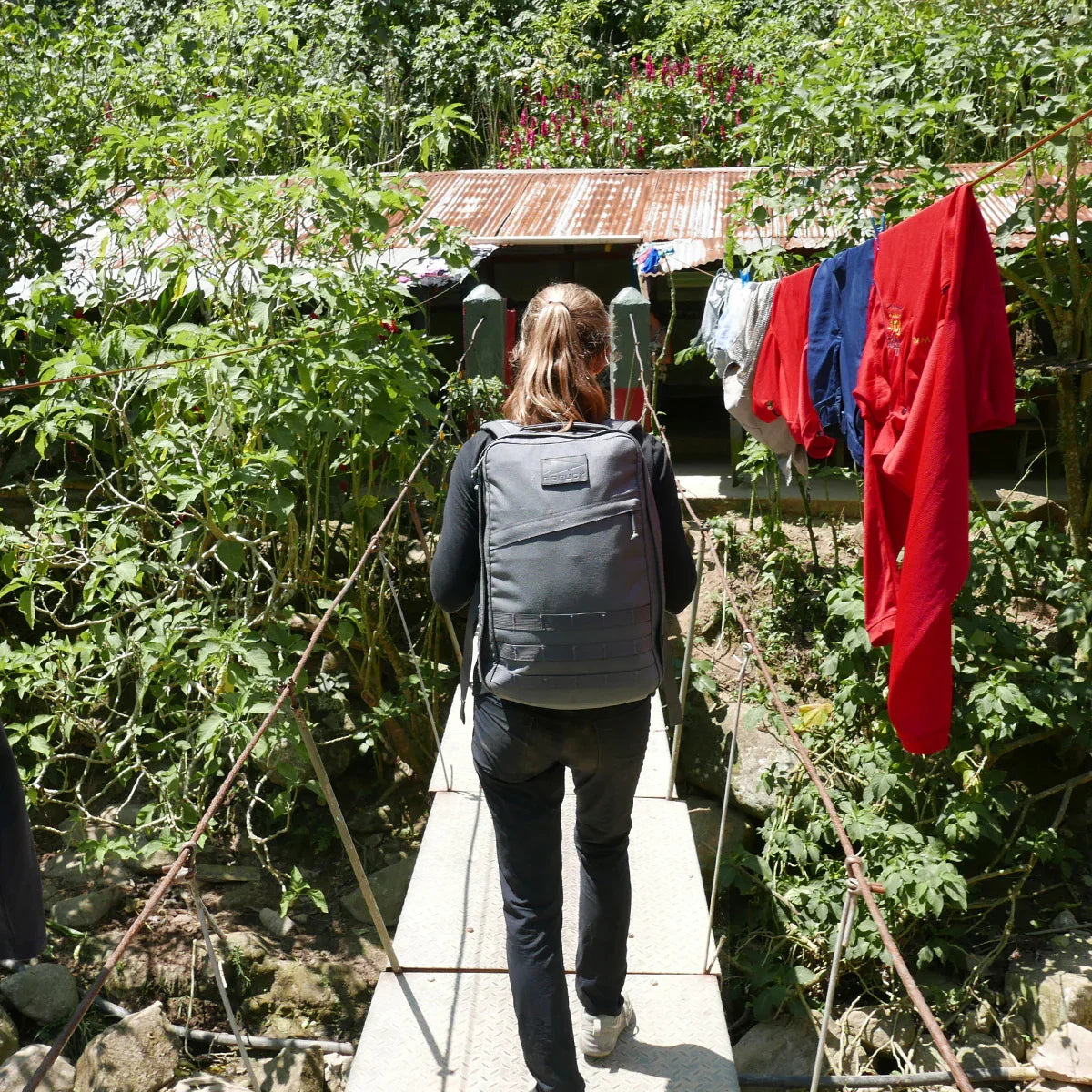 Person wearing GORUCK backpack crossing a footbridge outdoors, surrounded by greenery and laundry.