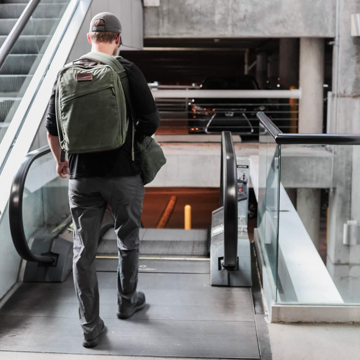 Man wearing olive green GORUCK backpack walking down escalator in urban parking garage