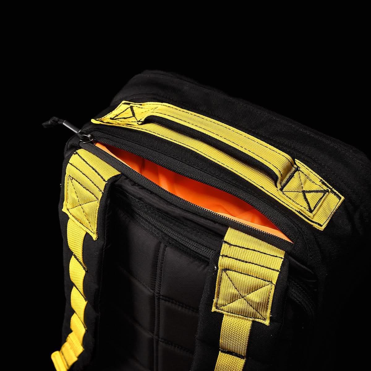 Close-up of black GORUCK backpack with yellow webbing handle and padded straps, partially unzipped showing orange interior