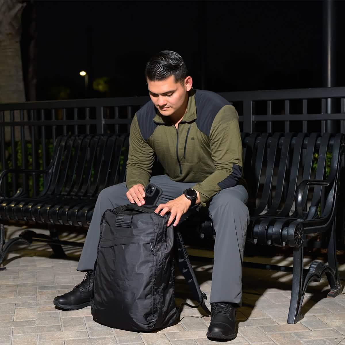 Man sitting on outdoor bench at night opening black GORUCK backpack, holding smartphone, wearing olive-green pullover and gray pants