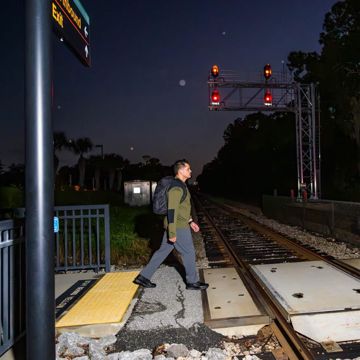 Man wearing black GORUCK backpack crossing railroad tracks at night with red signal lights