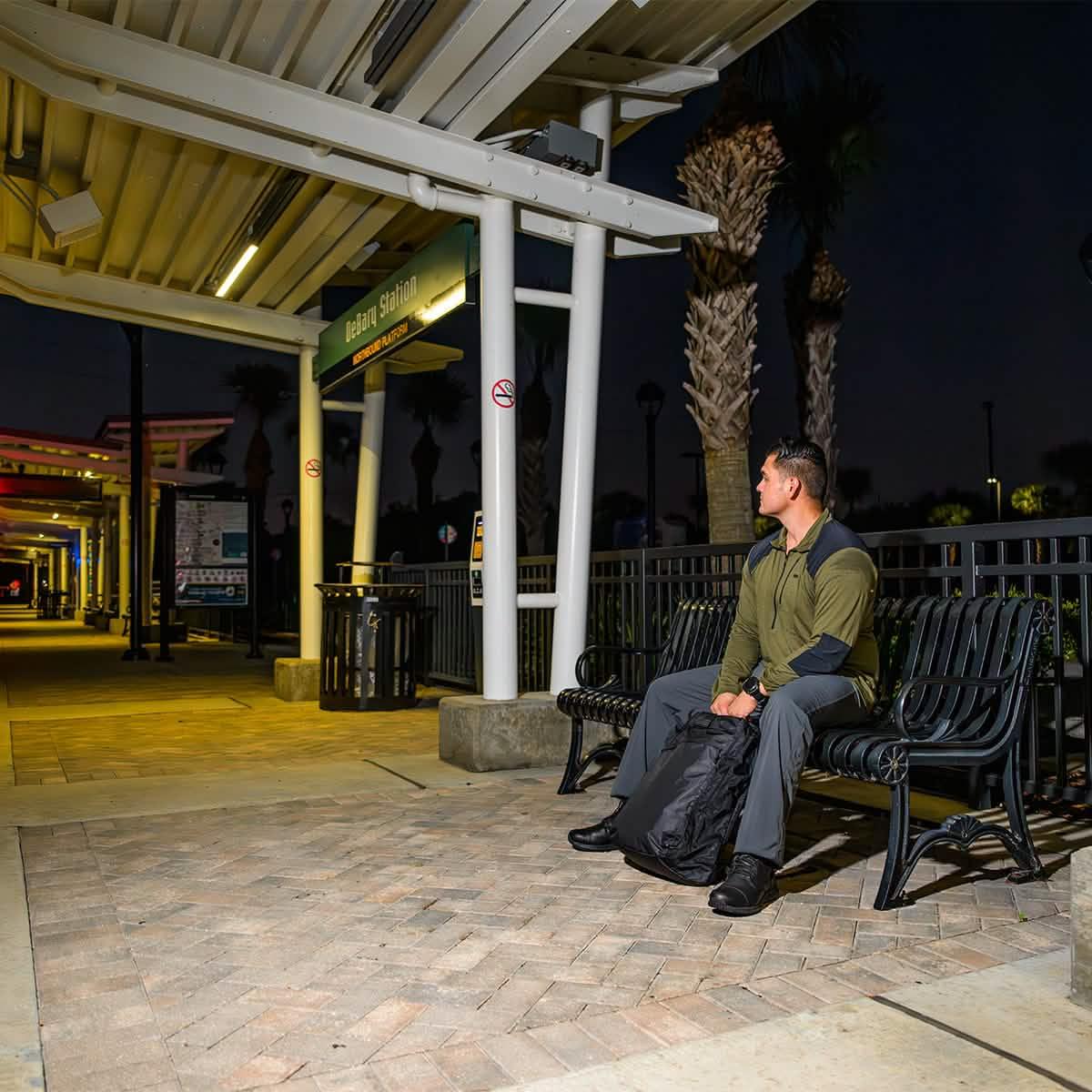Man in olive jacket sitting on a bench at night with black GORUCK backpack at DeBary station platform