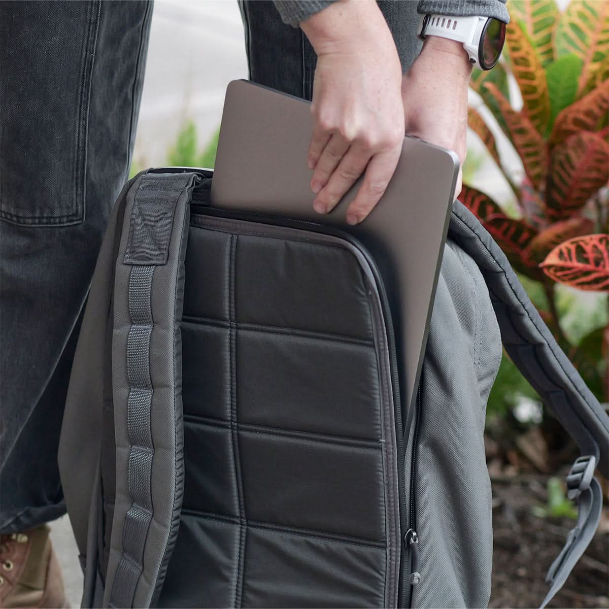 A person places a laptop into the bombproof compartment of a gray GR1 USA - Cordura rucksack by GORUCK, beside colorful plants, featuring Special Forces grade durability.