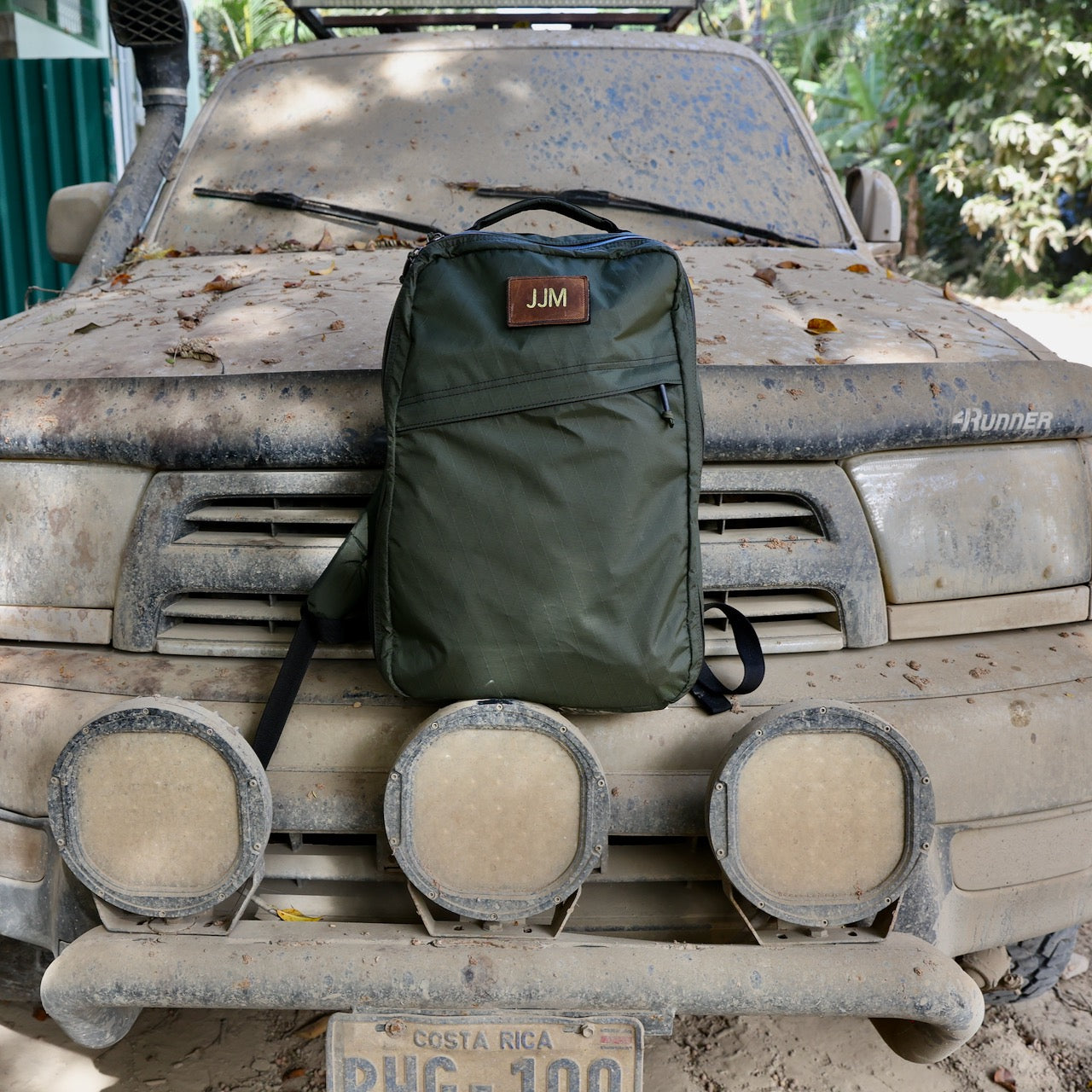 A green backpack featuring a Patch - Custom Leather Monogram sits on the dusty hood of a dirt-covered truck with Costa Rica plates.