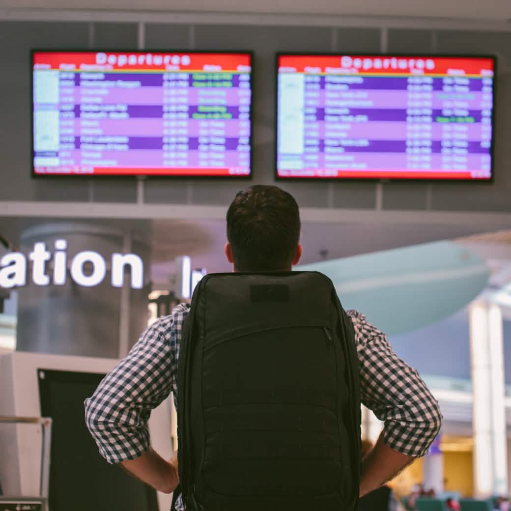 A traveler with a GORUCK GR2 backpack, designed to comply with carry-on regulations, stands before the departure screens at an airport, glancing up to check flight information. The screens show various flight destinations and times in the lively terminal.