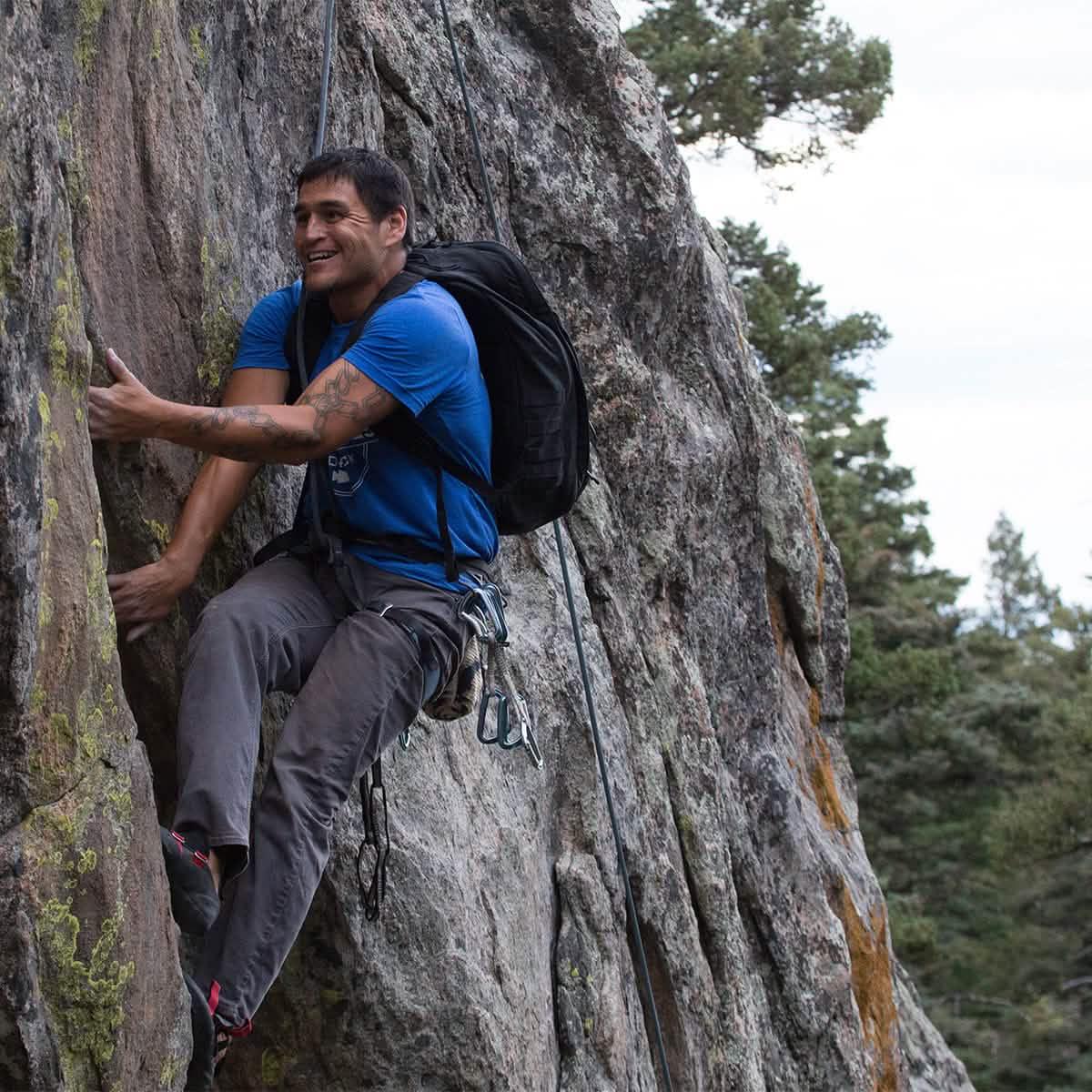 Man rock climbing outdoors wearing a blue shirt and black backpack with safety gear on rugged grey rock face