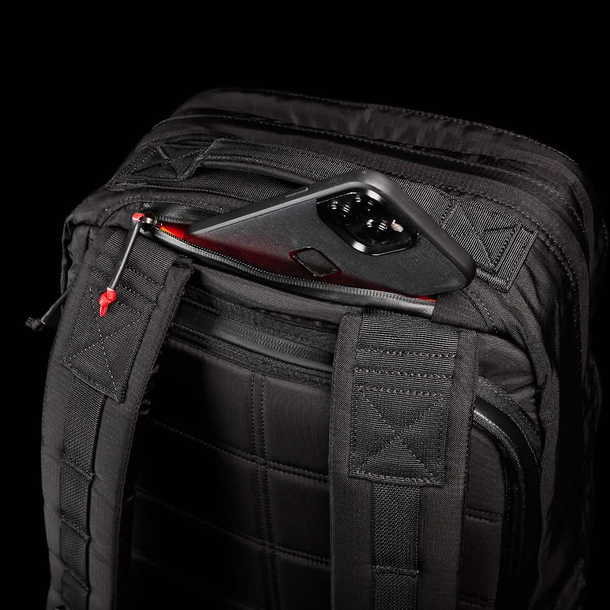 Introducing the GR2 - Ripstop ROBIC® by GORUCK, a stylish black rucksack complete with quilted straps and a compact external pocket that accommodates a smartphone in a red and black case. Crafted for ease of use, this water-resistant travel bag stands out against a solid black background.
