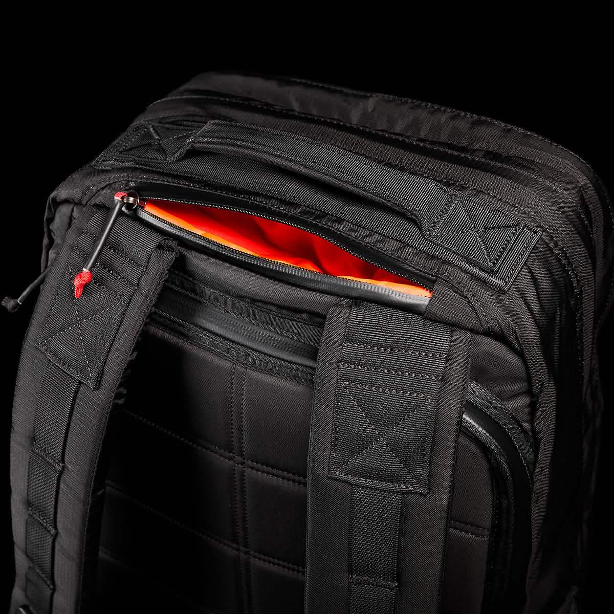 A close-up of the GR2 - Ripstop ROBIC® backpack by GORUCK showcases its black exterior with padded straps and a partially open top zipper pocket, revealing a bright orange interior. The textured, water-resistant fabric and stitching details of the rucksack stand out against a dark background.