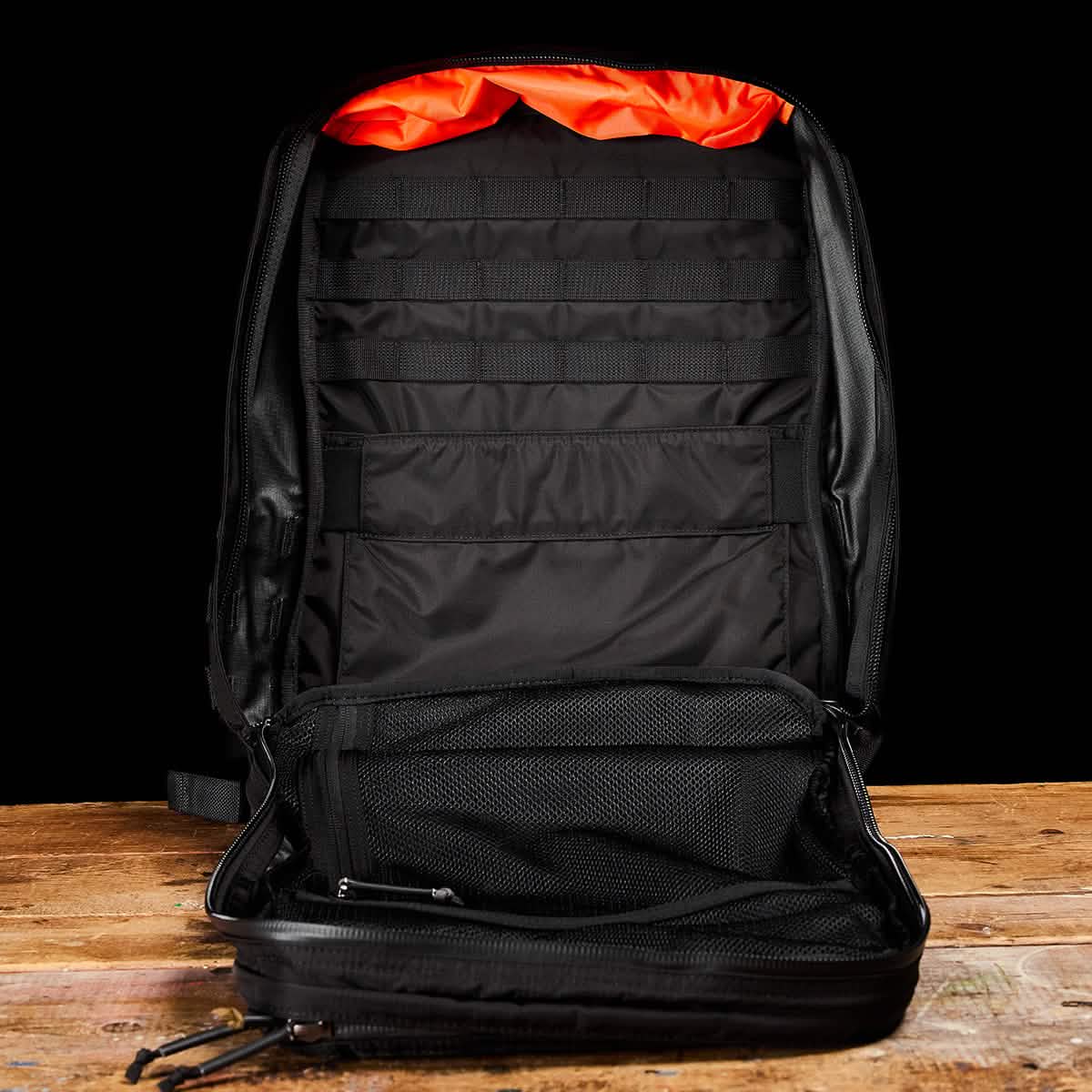 The open GR2 - Ripstop ROBIC® from GORUCK rests on a wooden surface, showcasing its numerous compartments and pockets. The interior features an orange fabric lining at the top. This unzipped, water-resistant travel bag is empty, highlighting its organized storage options.