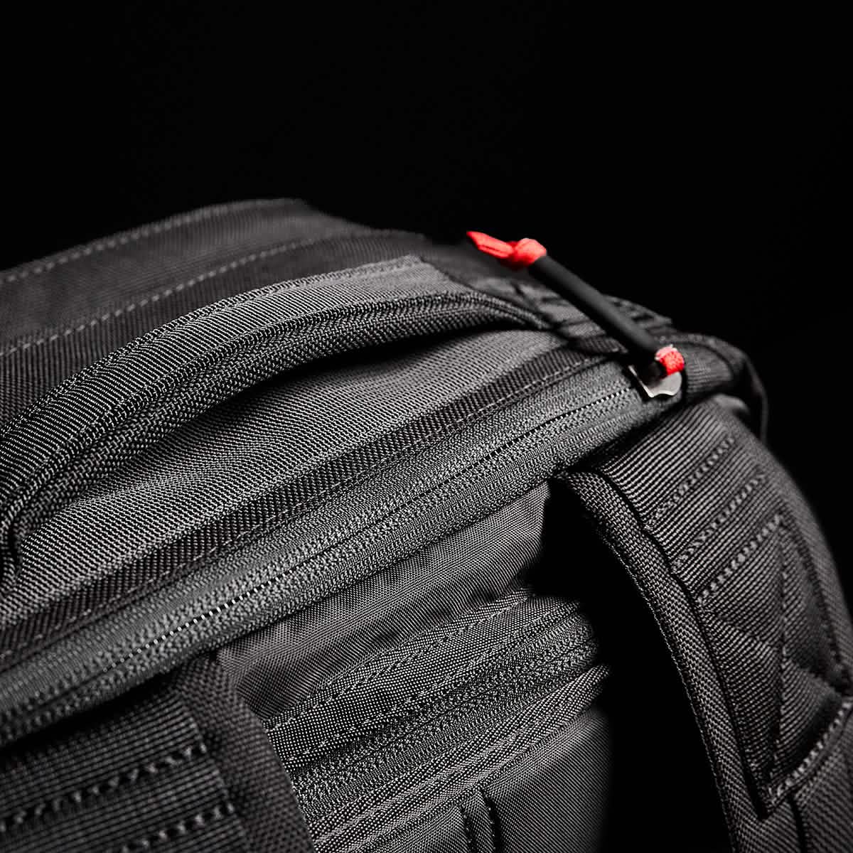 A close-up of a black zippered bag, echoing the robust design of the GORUCK GR2 - Cordura used by Special Forces, highlights vibrant red pull tabs set against a dark backdrop. The material looks durable, with clearly visible stitching and seams.