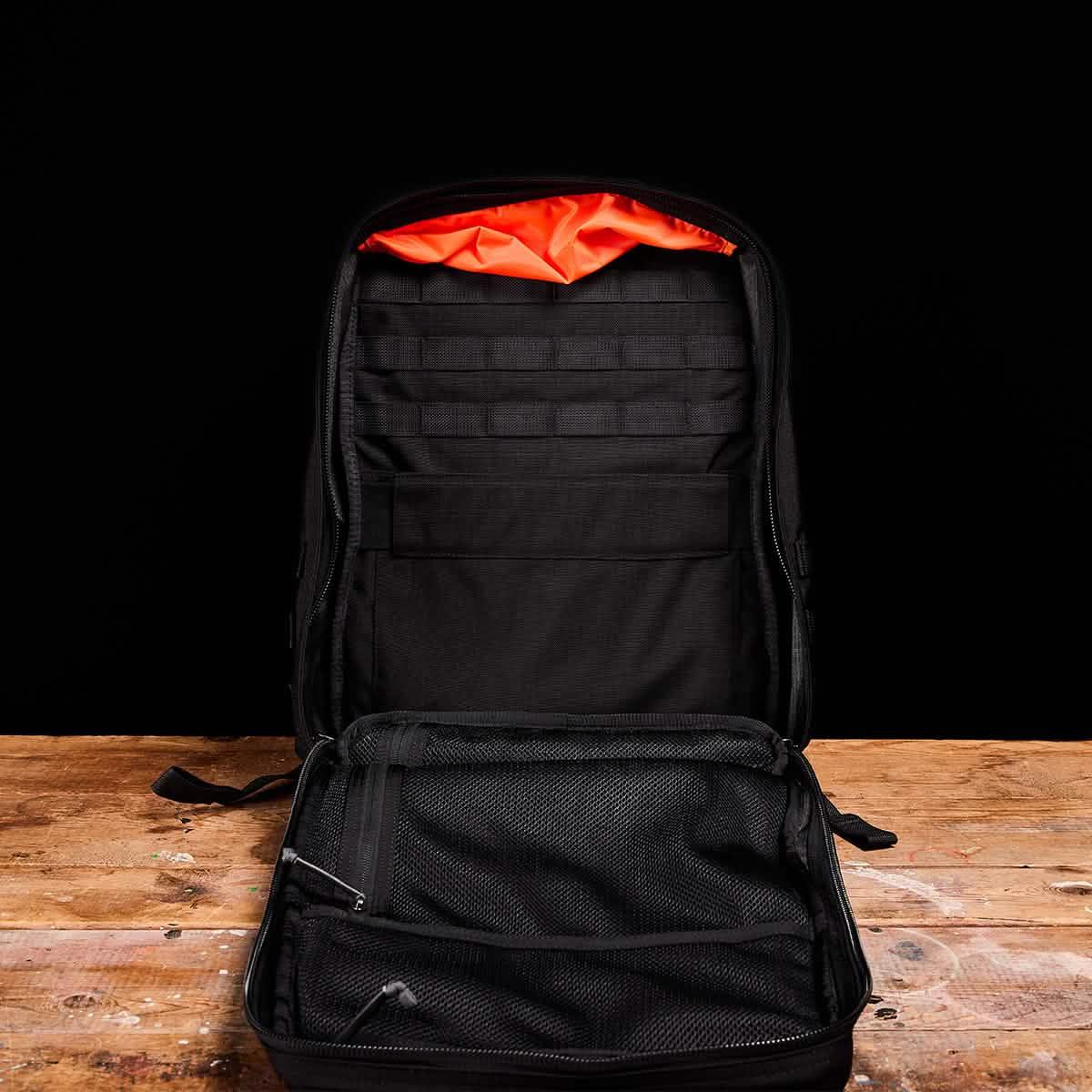 Open black tactical backpack with orange interior, MOLLE webbing, and mesh zippered compartments on wooden surface