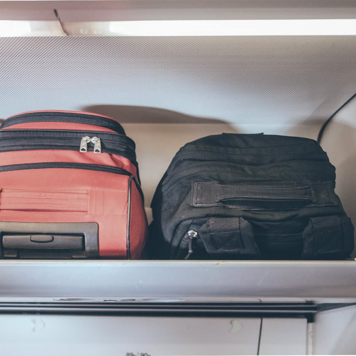 Two durable travel bags, one red with wheels and one black tactical style, stored on an overhead shelf