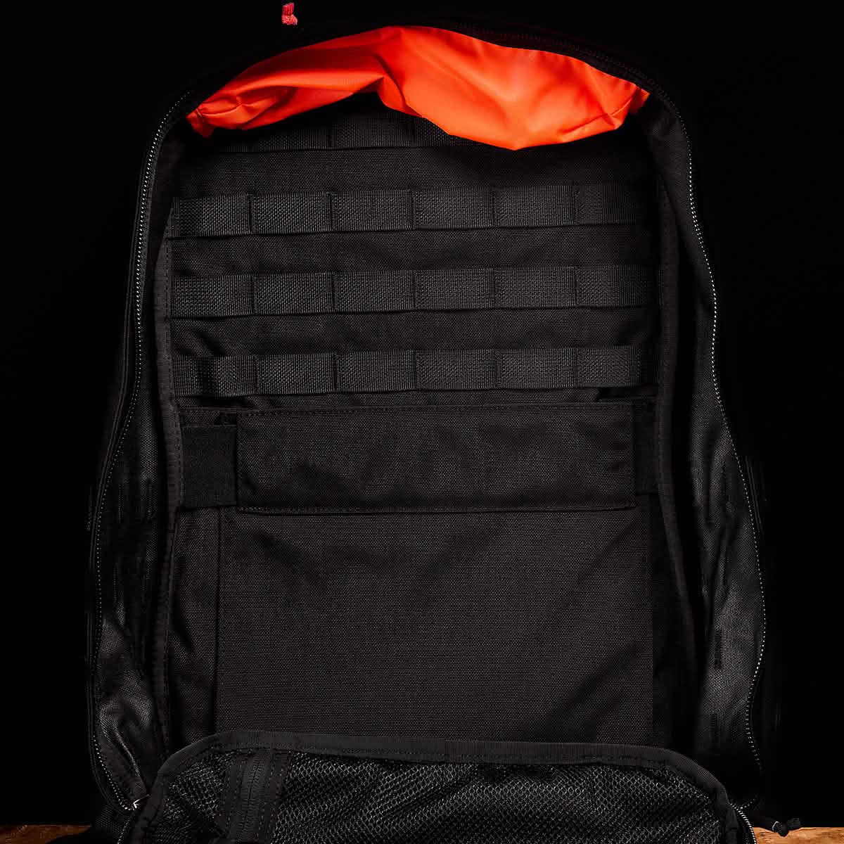The GR2 by GORUCK, a black ruck designed for Special Forces use, is set against a matching background and features an eye-catching orange interior lining. It resembles a travel bag with multiple storage compartments, including mesh pockets and webbing for optimal organization.