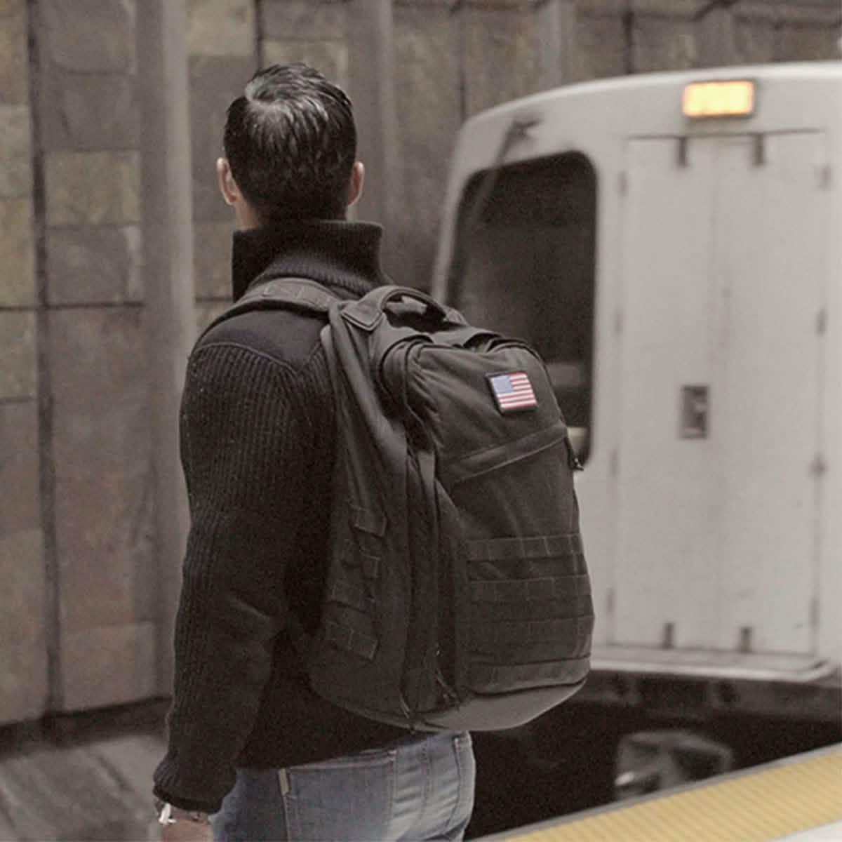 Man wearing black GORUCK backpack with American flag patch standing on subway platform