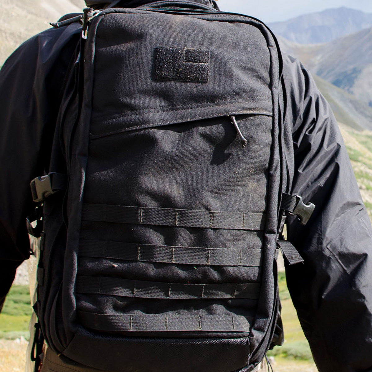 A person wears the GR2 USA Throwback, a large black travel backpack crafted from rugged 1000D Cordura, outdoors in the mountains.