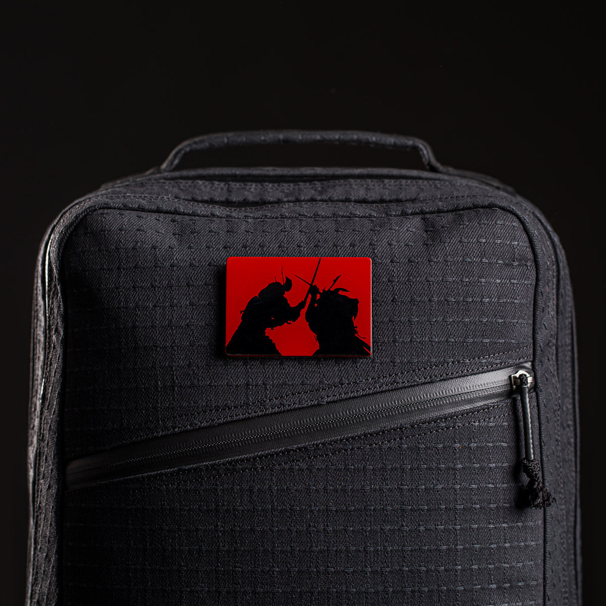 A square pin featuring two samurai silhouettes on red, inspired by the GR2 x Carryology - GRXC2 Samurai 2.0, is attached to the front pocket of a black textured bag.