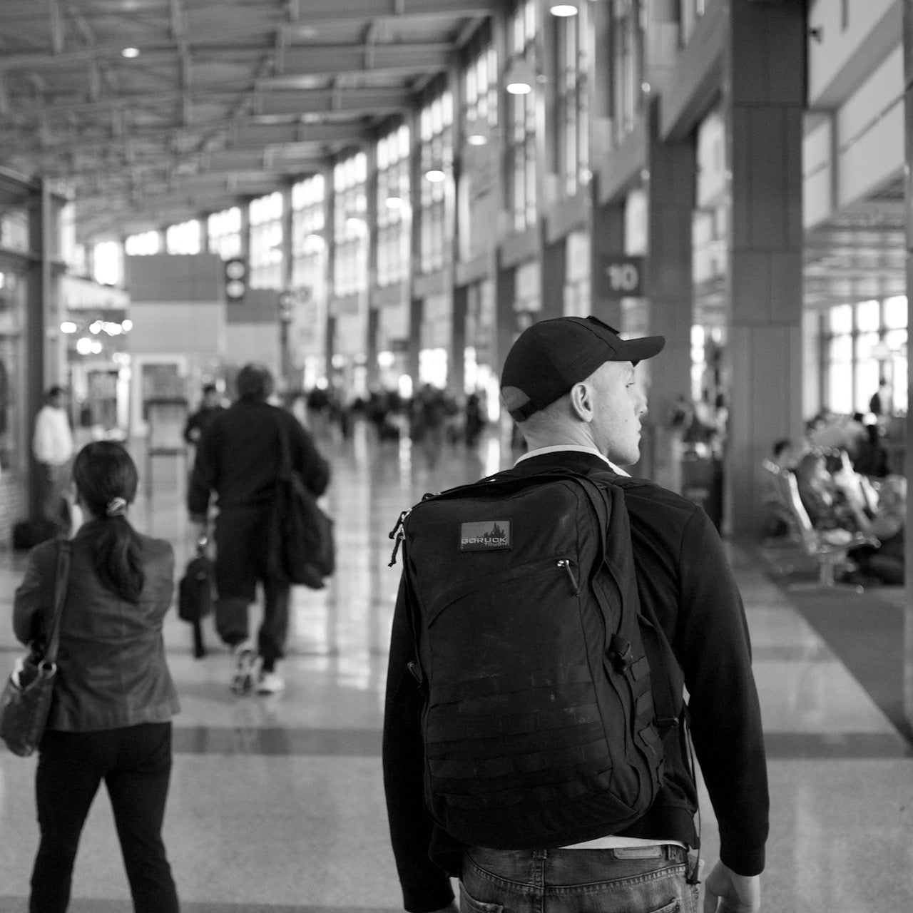 A man wearing a cap carries the GR2 USA - Cordura travel backpack while walking through a busy airport terminal in black and white.
