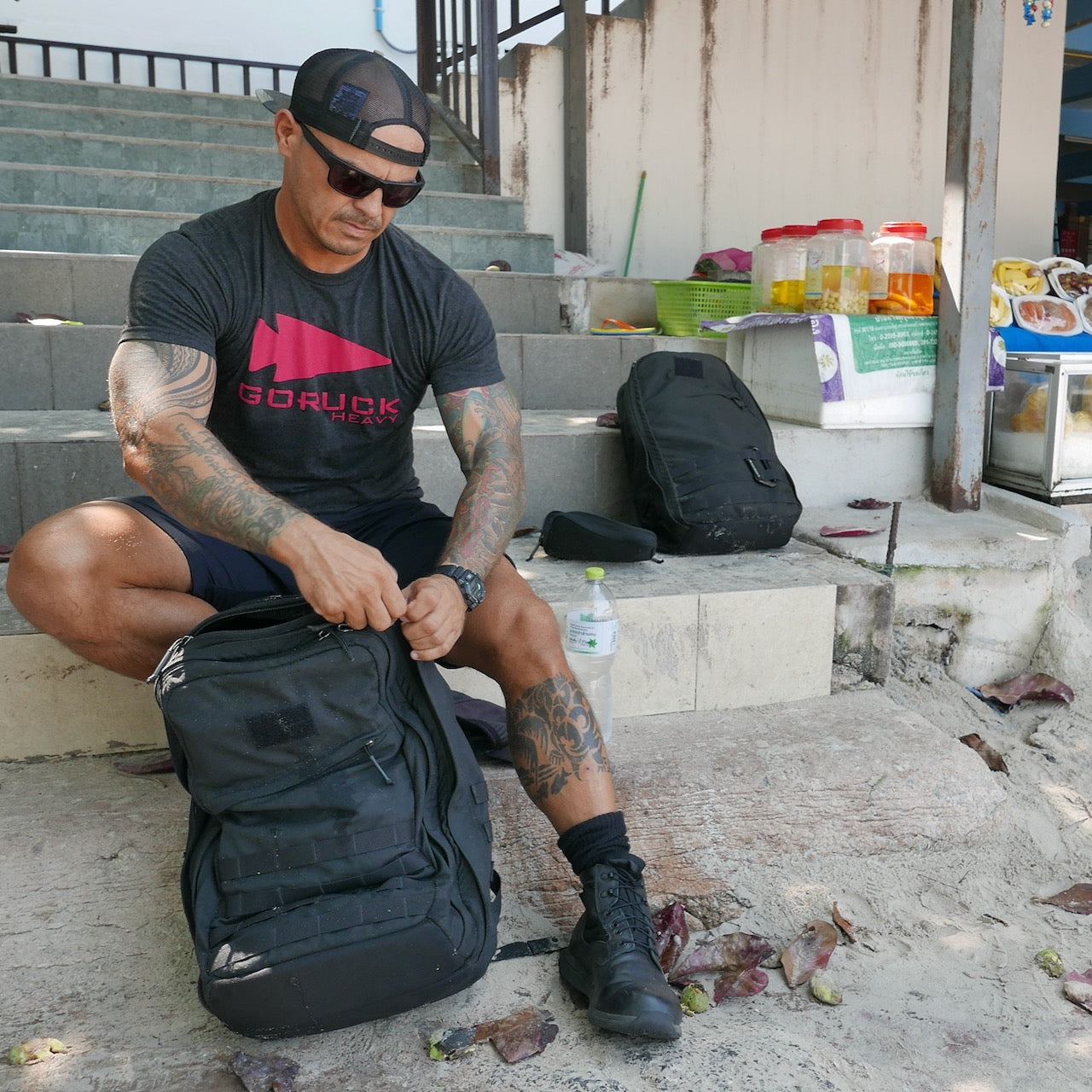 A man in a black t-shirt and boots sits on steps, zipping his GR2 USA - Cordura travel backpack, with food jars and another bag nearby.