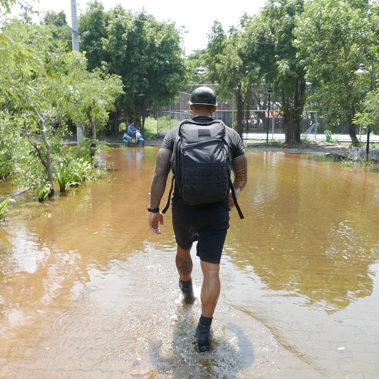 A man carrying the GR2 USA – Cordura travel backpack walks through a flooded path lined with trees on a sunny day.