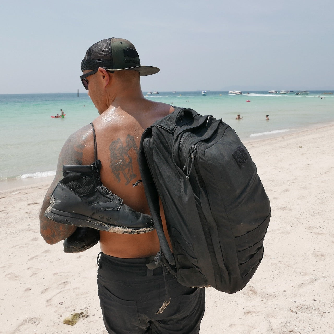 A man on a sandy beach carries the GR2 USA - Cordura travel backpack and boots, with people swimming and boats visible in the background.