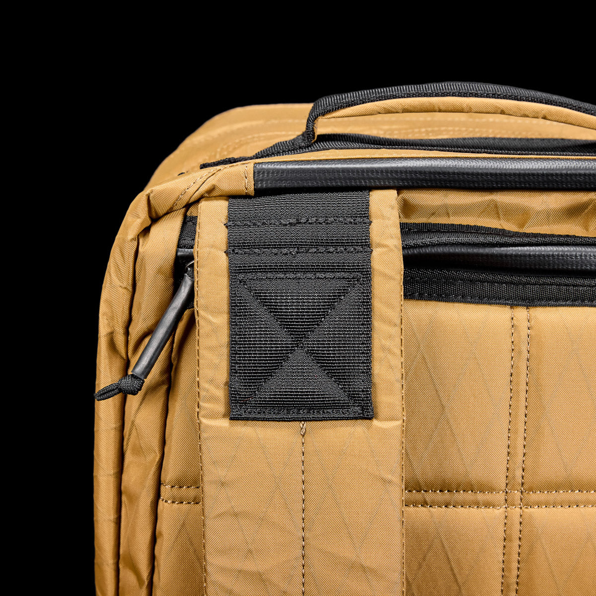 Close-up of a tan quilted bag with black zippers and handle against a black background.