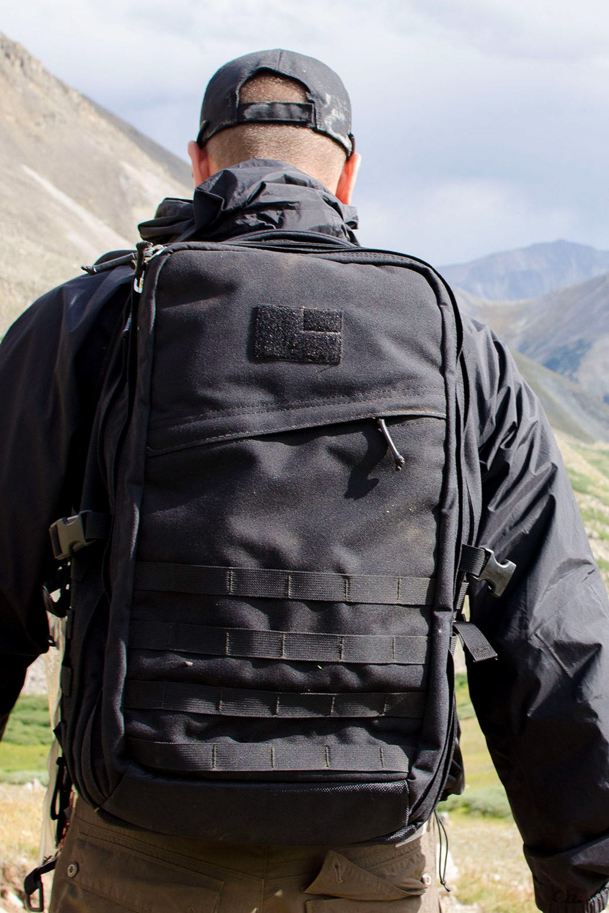 Person wearing a black jacket and backpack, standing outdoors with mountains in the background.
