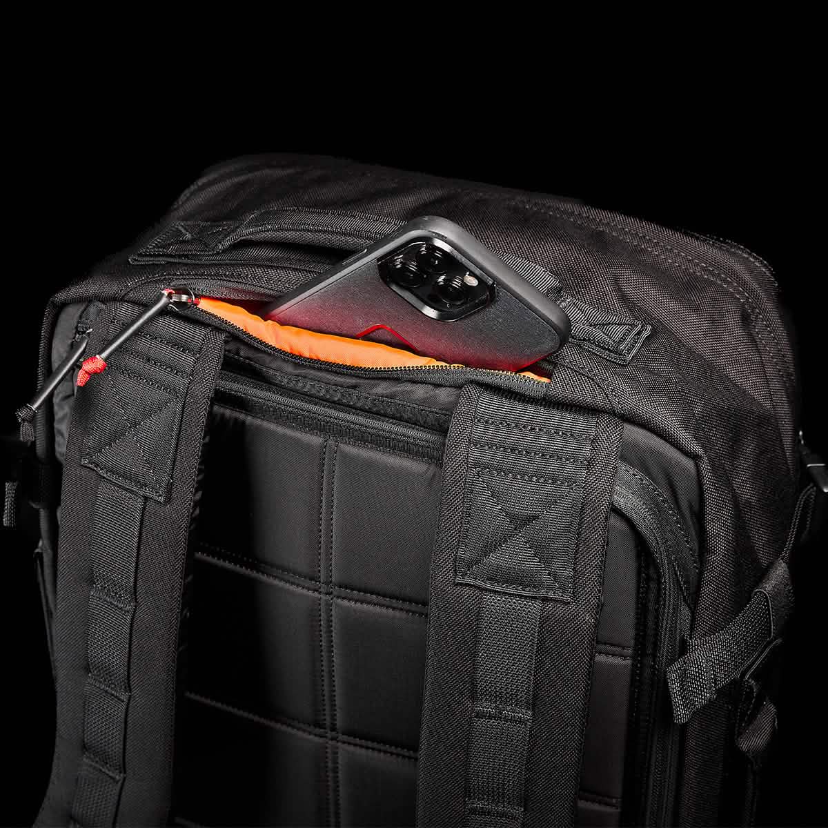 A GR3 - Cordura carry-on rucksack from GORUCK rests on its side, with a smartphone encased in metallic peeking out of an open pocket. This sleek black backpack showcases water-resistant material, padded straps, and a quilted GR3® design on the back.