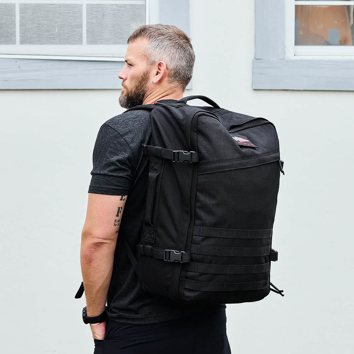 A man in a black T-shirt stands sideways, carrying the water-resistant GR3 - Cordura rucksack from GORUCK. He's positioned in front of a light-colored wall with two windows visible.