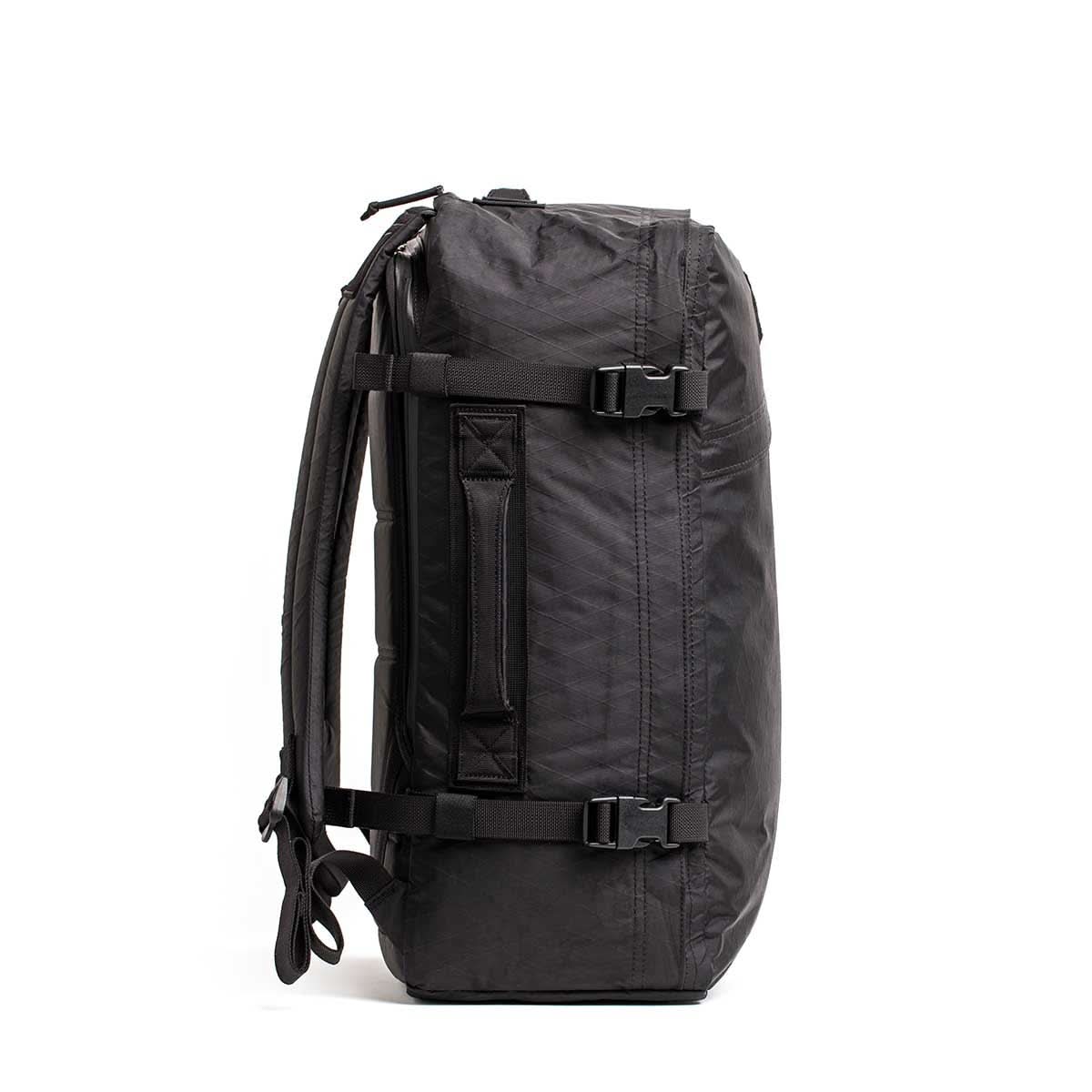 Side view of the GR3 - X-PAC, a black waterproof carry-on backpack with straps and buckles, standing upright against a white background.