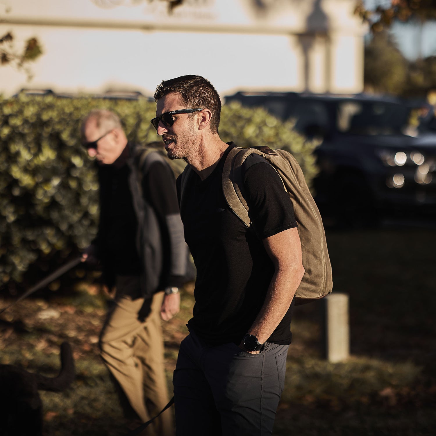A man in sunglasses and a backpack sports the Men’s Commando Tee - Merino Wool, featuring reinforced shoulders, while walking outdoors with another person visible in the sunny background.