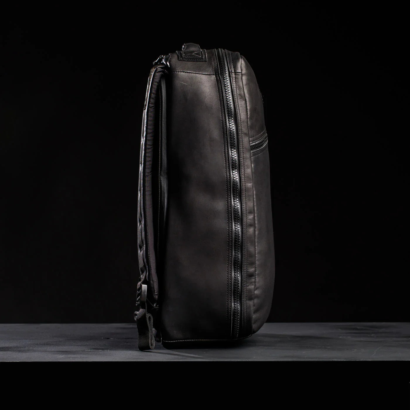 Side view of sleek black leather tactical rucksack with zippers on dark background