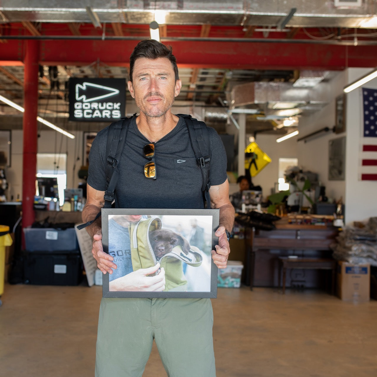 A man in a black shirt and backpack holds a framed photo inside an industrial-style room with red pipes, his GR MONSTER Rucksack - PREORDER slung over one shoulder.