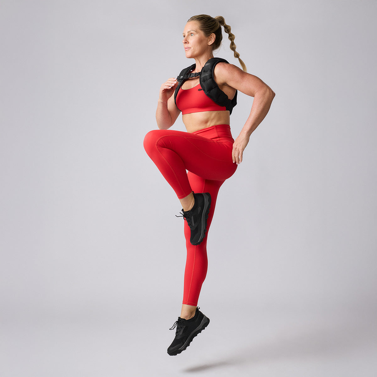Woman in red workout clothes and black shoes doing a high knee exercise with a weighted vest.