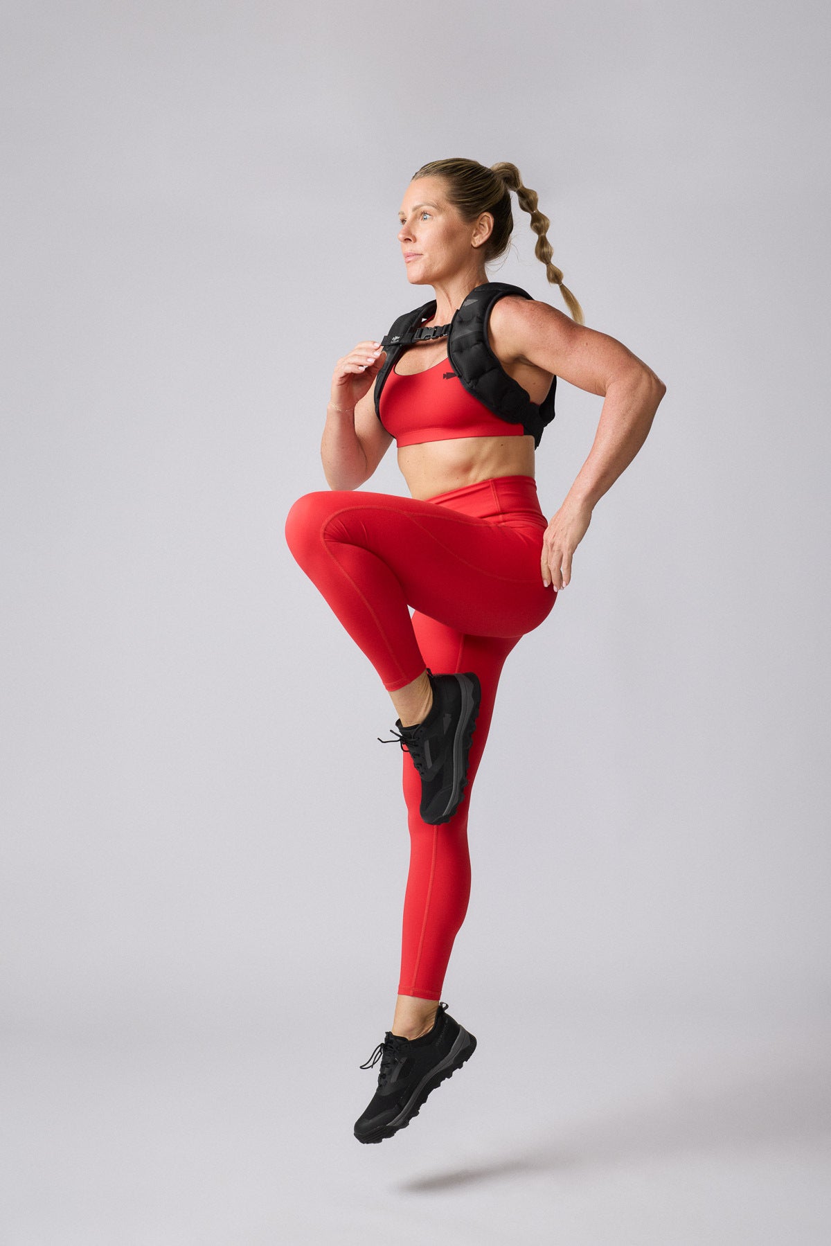 A woman in red squat-proof Training Leggings, made in the USA, wears a weighted vest while performing high knees against a plain background.