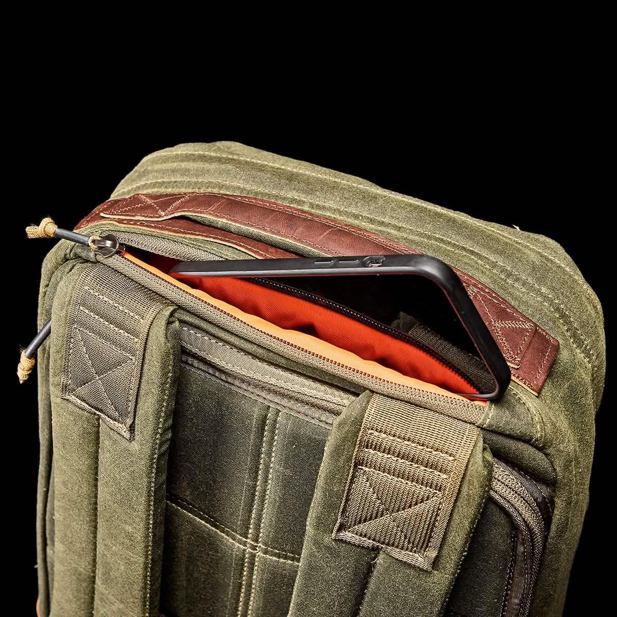 Close-up of olive green tactical rucksack with tan strap and partially open zipper showing an orange interior and phone pocket