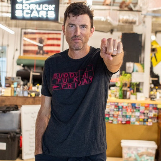 A man wearing the Buddy FCKR Friday Tee - Tri-BlendX stands indoors, pointing at the camera with a “GORUCK SCARS” sign behind him—showcasing his GORUCK pride and readiness to pay it forward.