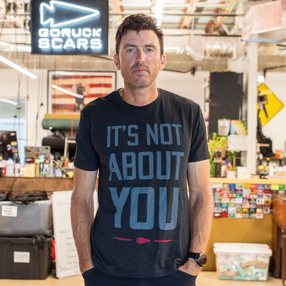 A man wearing the “It’s Not About You - Tri-BlendX - Unisex” shirt stands in an office with industrial decor and signs. This premium Tri-BlendX shirt, printed in the USA, combines style and comfort for everyday wear.