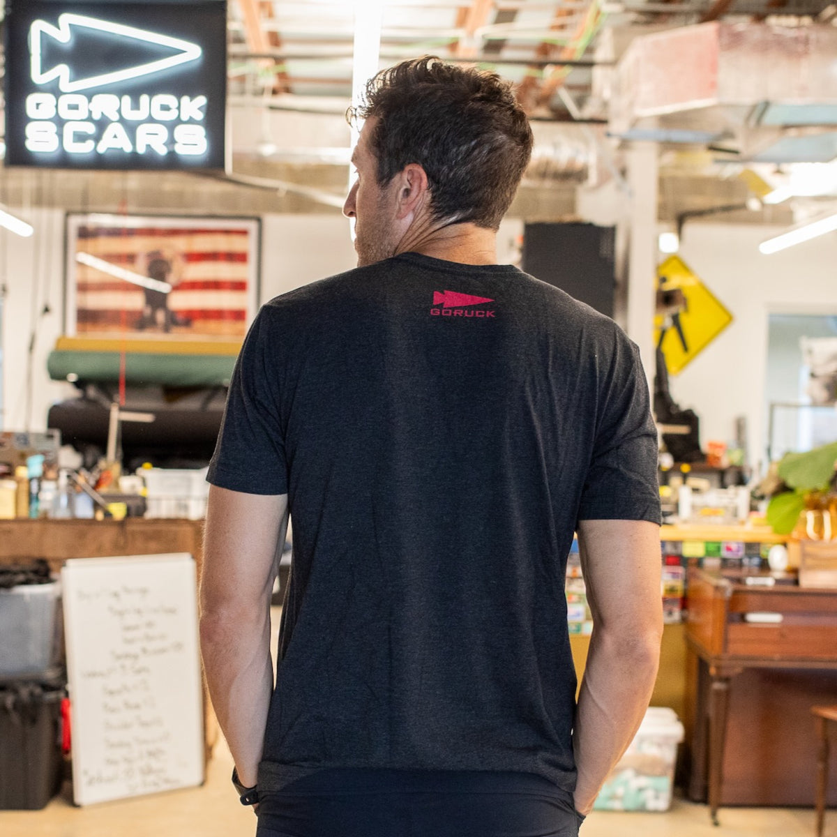 A man stands indoors, facing away, wearing the “It’s Not About You - Tri-BlendX - Unisex” t-shirt in black—crafted from premium Tri-BlendX fabric and printed in the USA—with equipment and signs visible in the background.