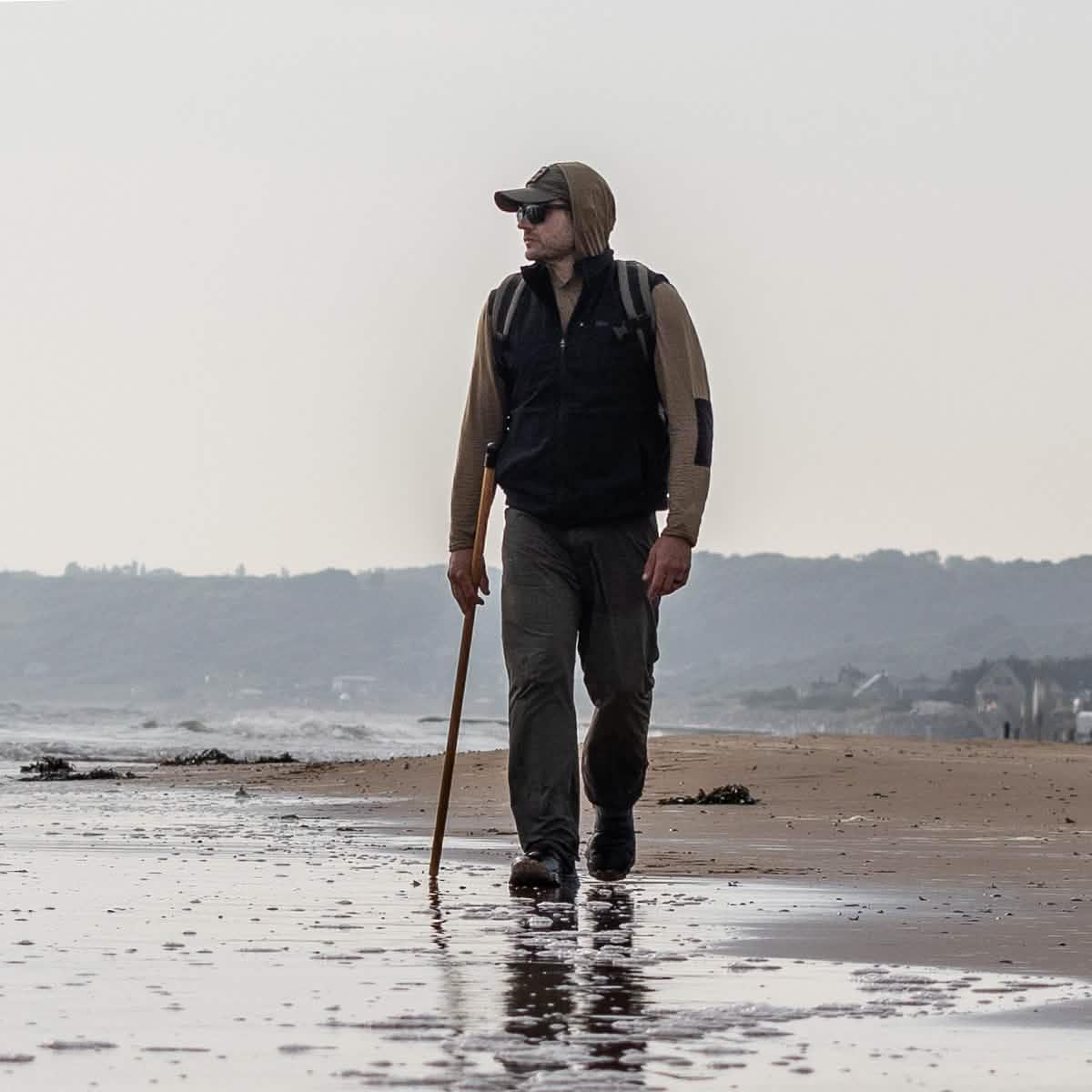 Man in GORUCK rucking gear hiking with a stick along a beach shoreline