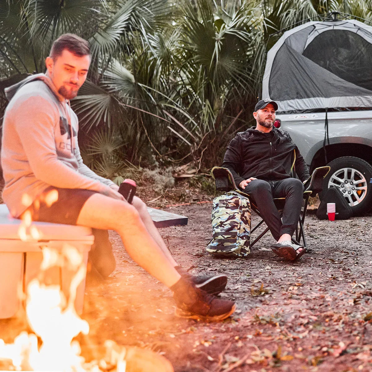 Two men relaxing by a campfire with backpack, camping chair, and truck with rooftop tent in forest setting