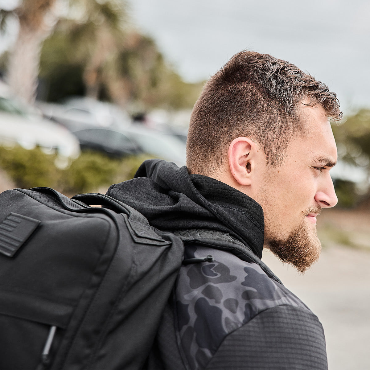 A man with short hair and a beard is outdoors wearing a Men’s Full Zip - Polartec Grid Fleece by GORUCK, and carrying a black backpack.